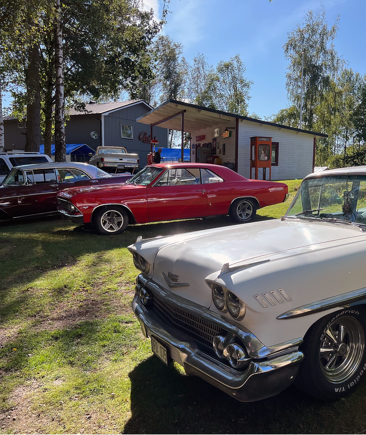 GM-bilar från olika decennier parkerade på en gräsmatta framför en gammal Texaco-mack med en orange telefonkiosk intill.